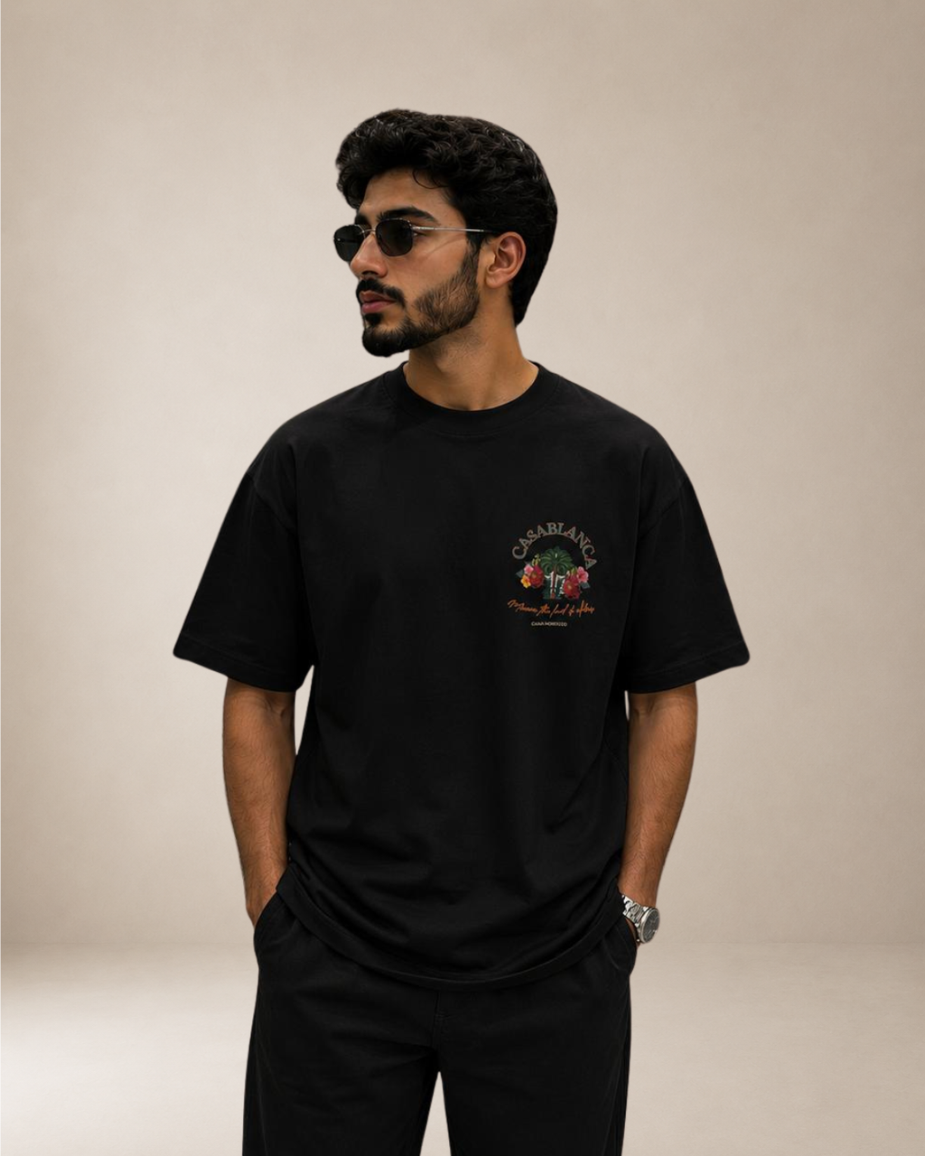 A man wearing a black short-sleeve t-shirt with a colorful graphic on the left chest, paired with black pants, set against a plain beige studio background.