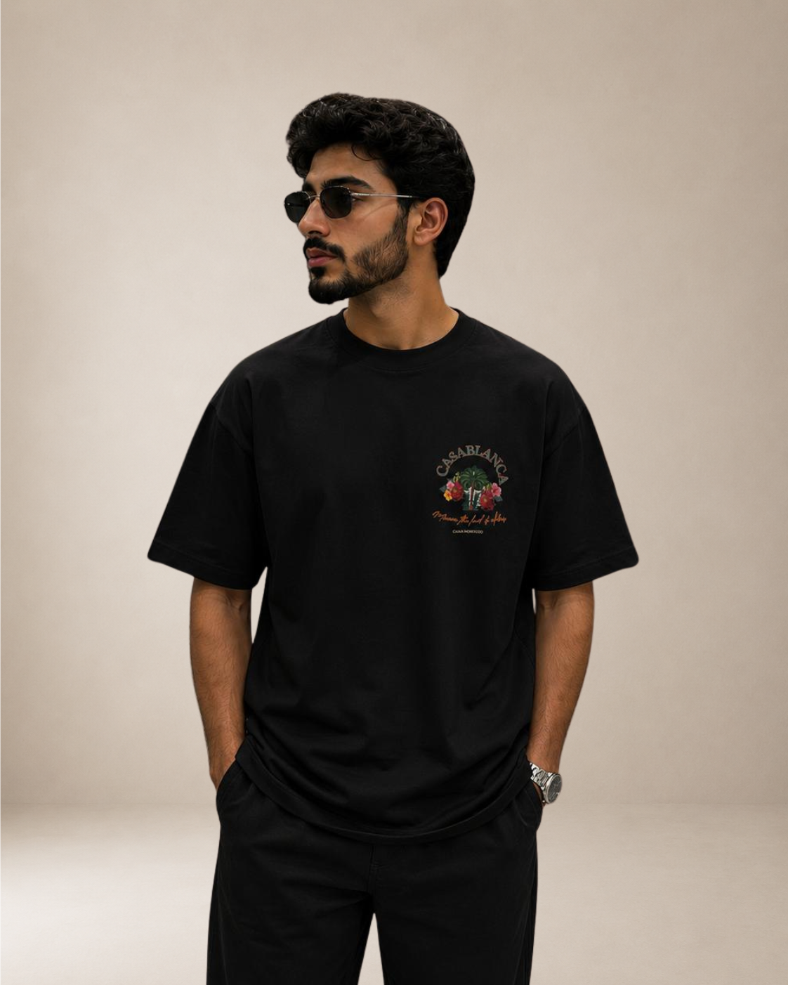 A man wearing a black short-sleeve t-shirt with a colorful graphic on the left chest, paired with black pants, set against a plain beige studio background.